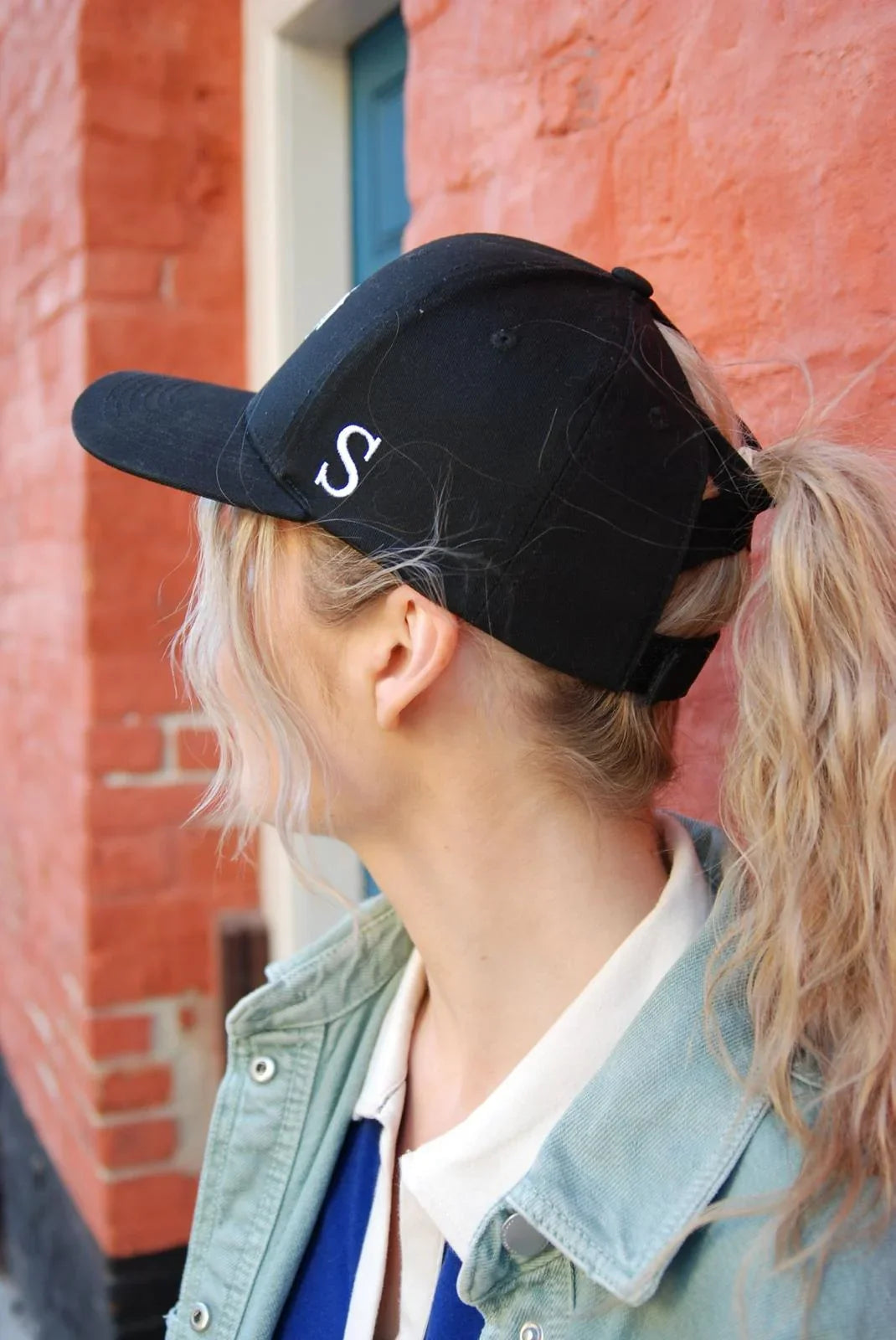 Person wearing a black ponytail baseball cap with a 'S' logo against a red brick wall.