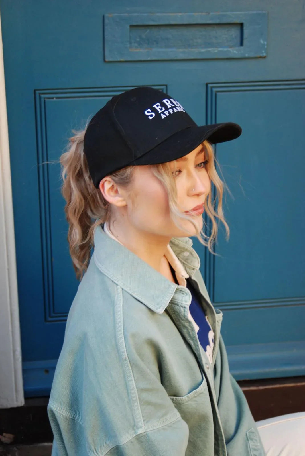 Person wearing a black ponytail baseball cap with 'SEREN Apparel' branding on the front in front of a blue door