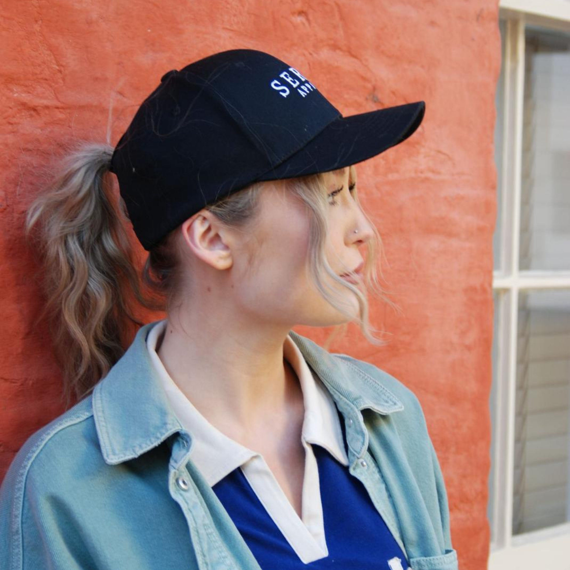 Person wearing a black ponytail baseball cap and denim jacket against a red wall.