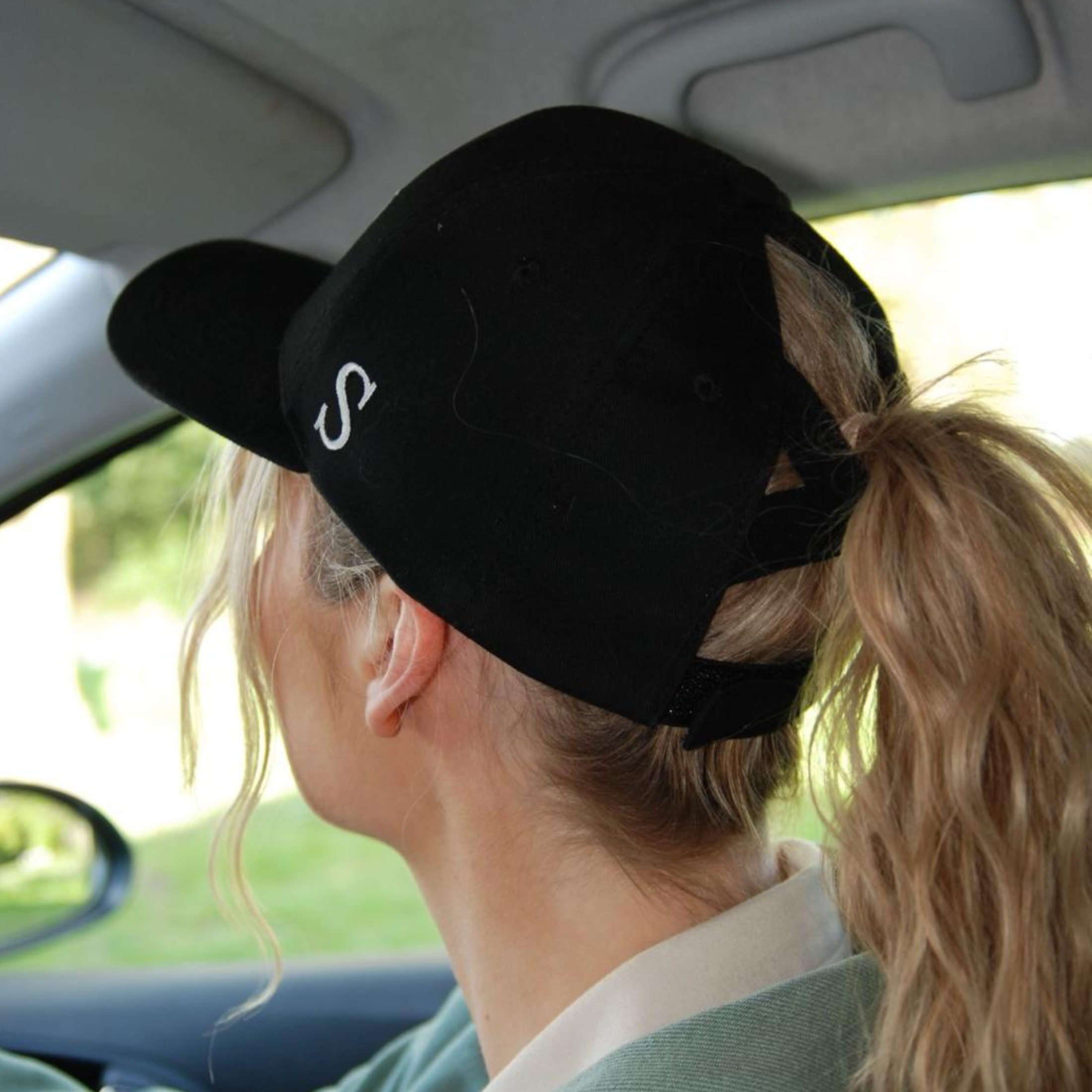 Person wearing a black ponytail baseball cap with a logo while sitting in a car.