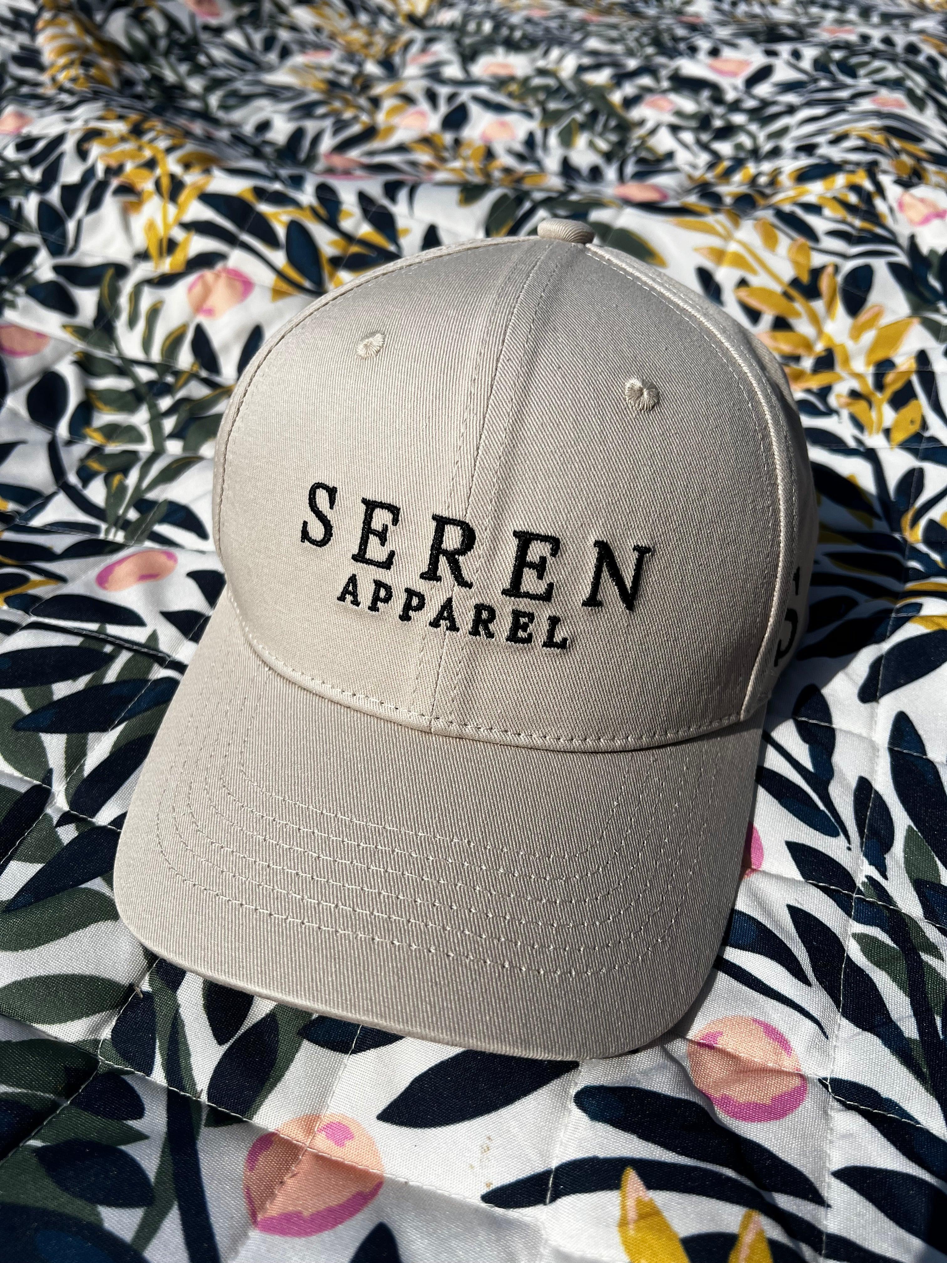 Beige ponytail baseball cap with 'Seren Apparel' logo on a floral fabric background