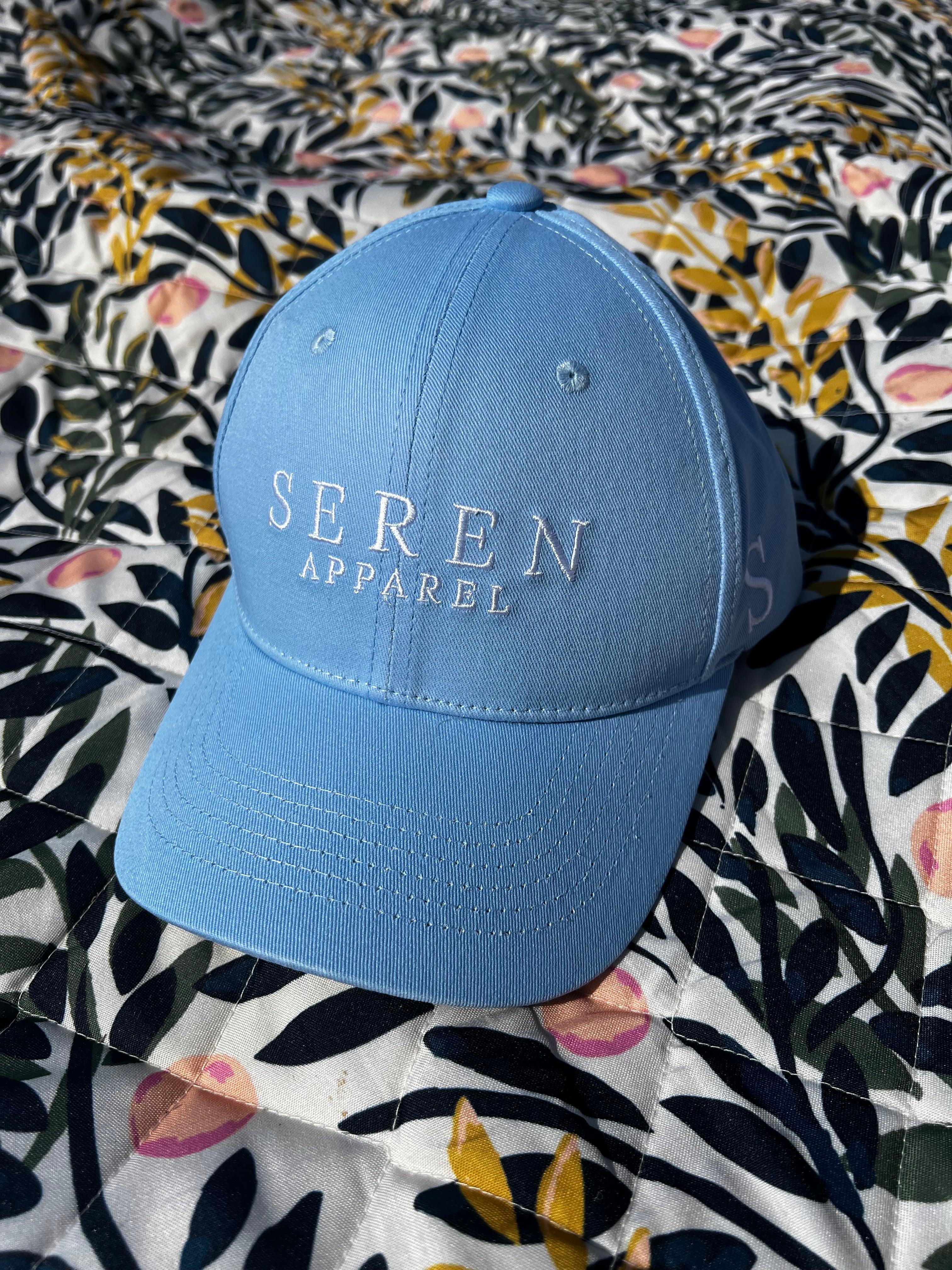 Light blue ponytail baseball cap with 'Seren Apparel' logo on a floral-patterned fabric background