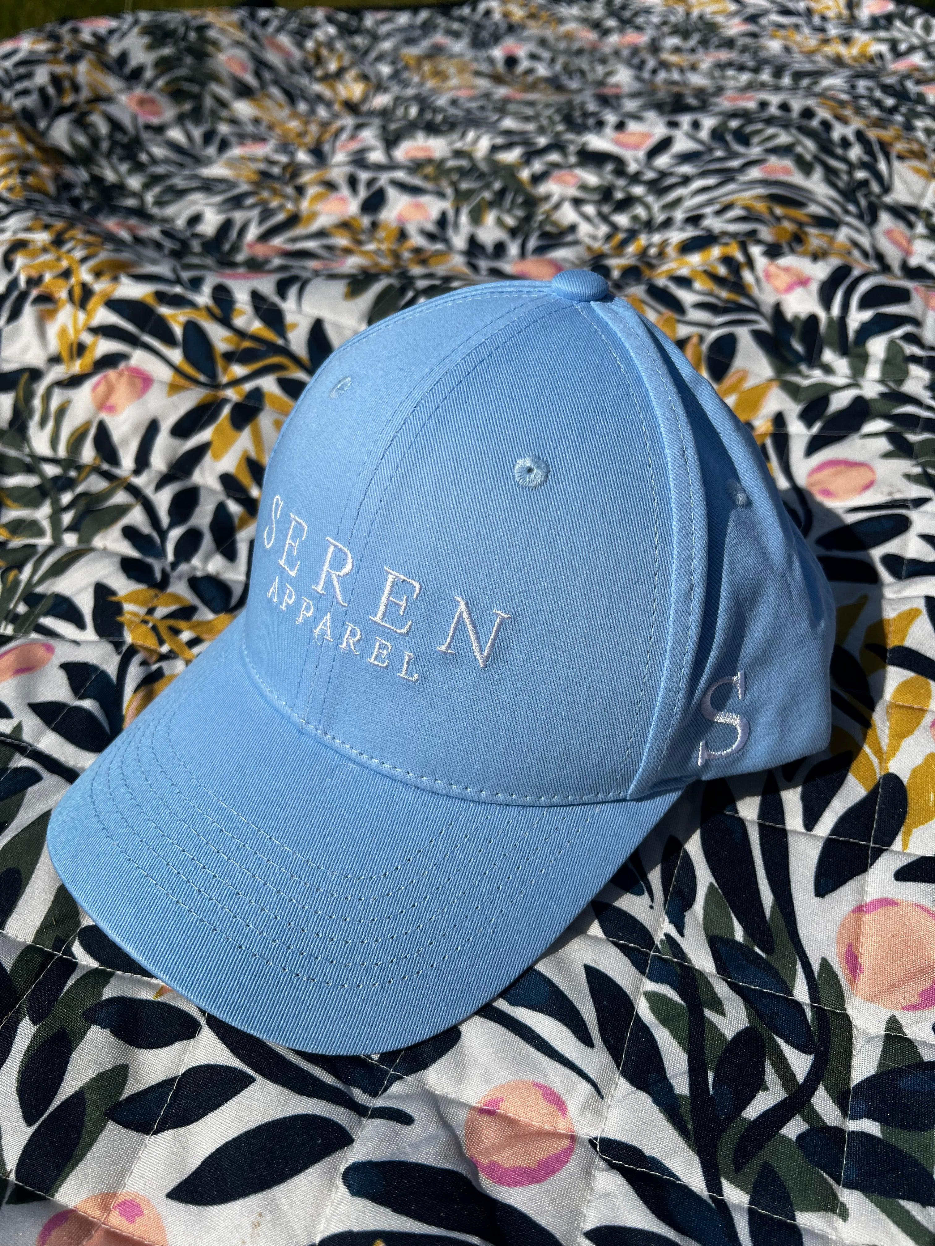 Light Blue ponytail baseball cap with 'SEREN APPAREL' logo on a floral fabric background