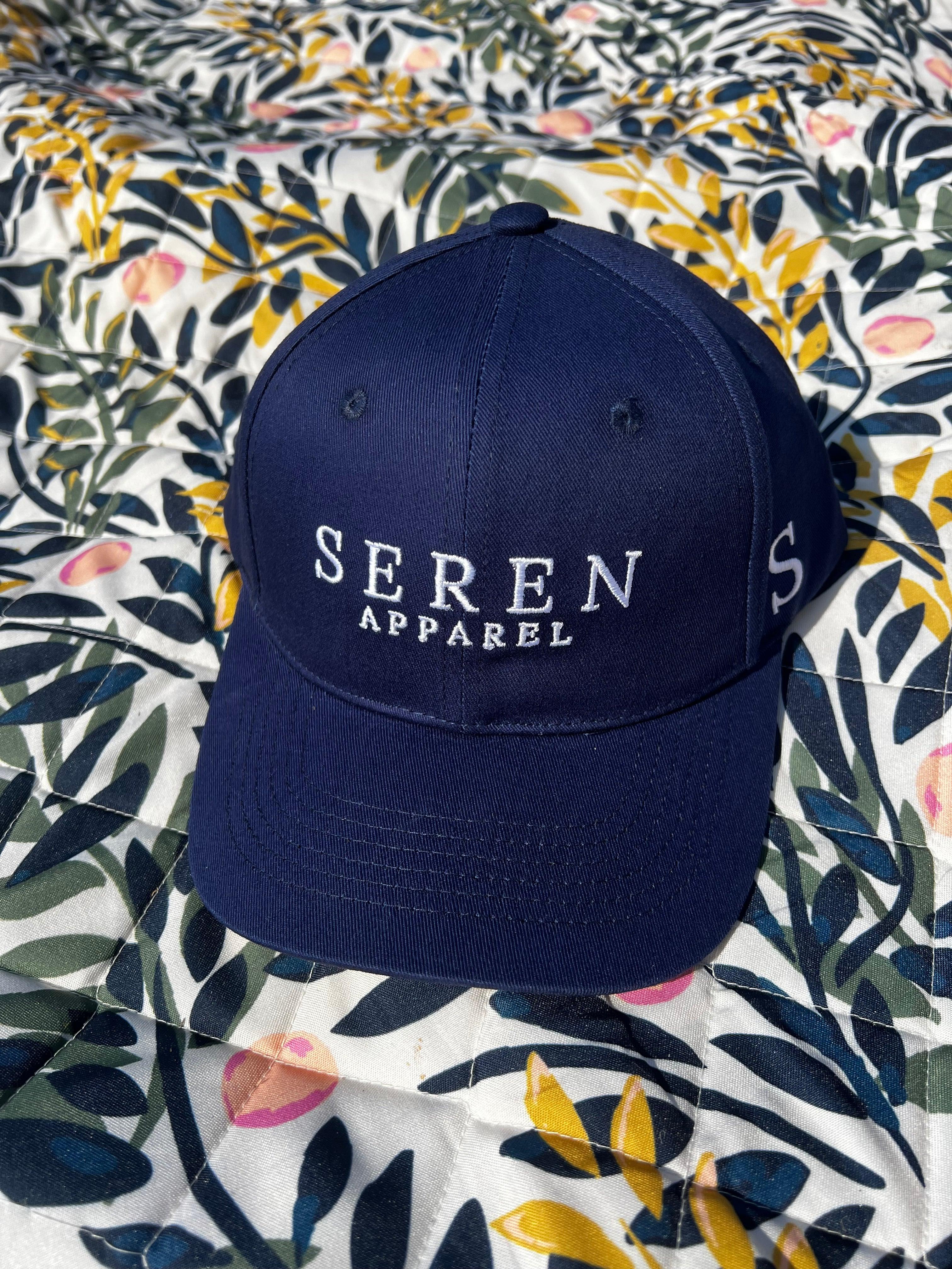 Navy blue ponytail baseball cap with 'Seren Apparel' logo on a floral fabric background